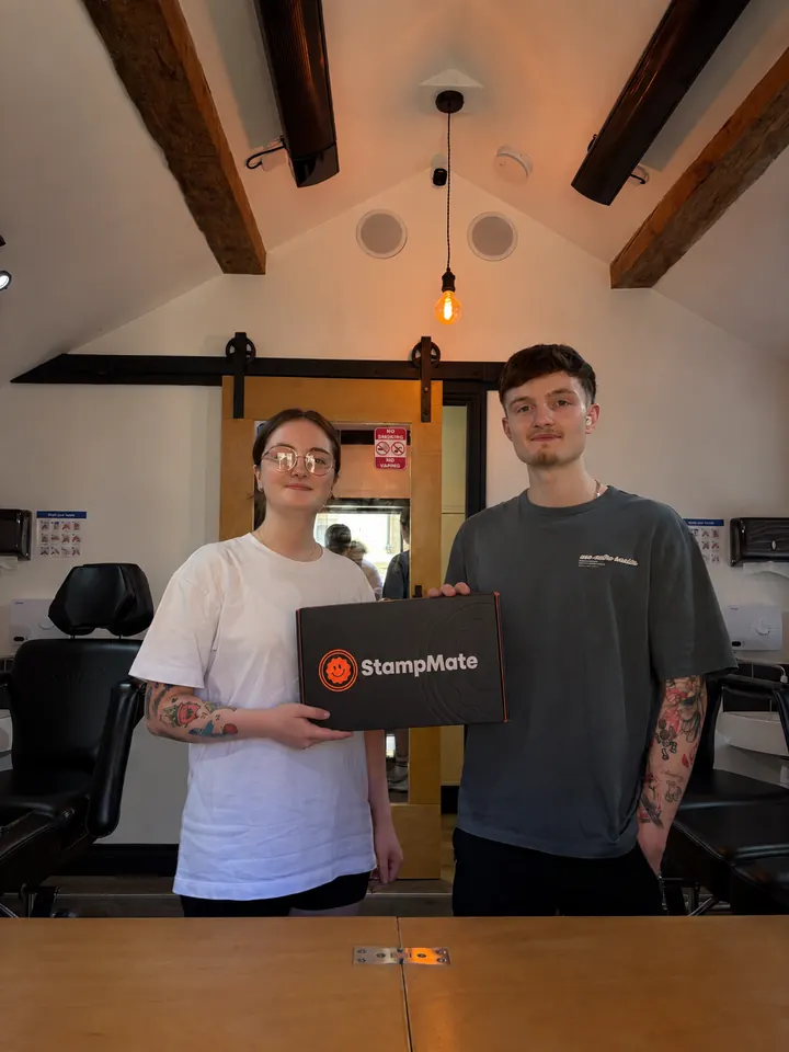 Two barbers holding a StampMate box in a shop with a barber chair